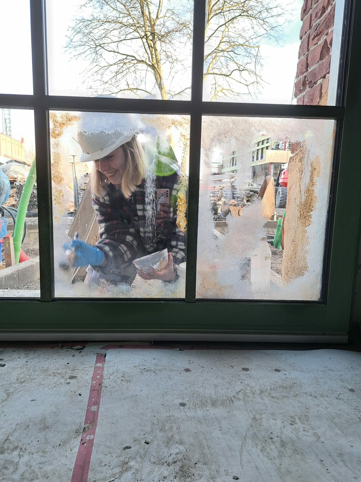 Femme souriante en casque de chantier appliquant une substance blanche sur une fenêtre vitrée.