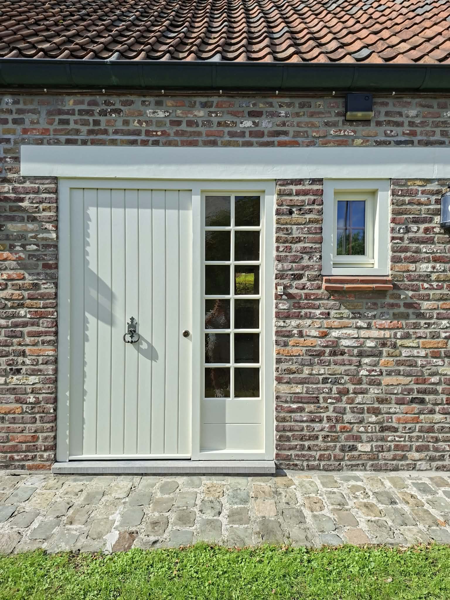 Porte crème sur mur de briques avec fenêtre à carreaux, toit de tuiles et pavés.