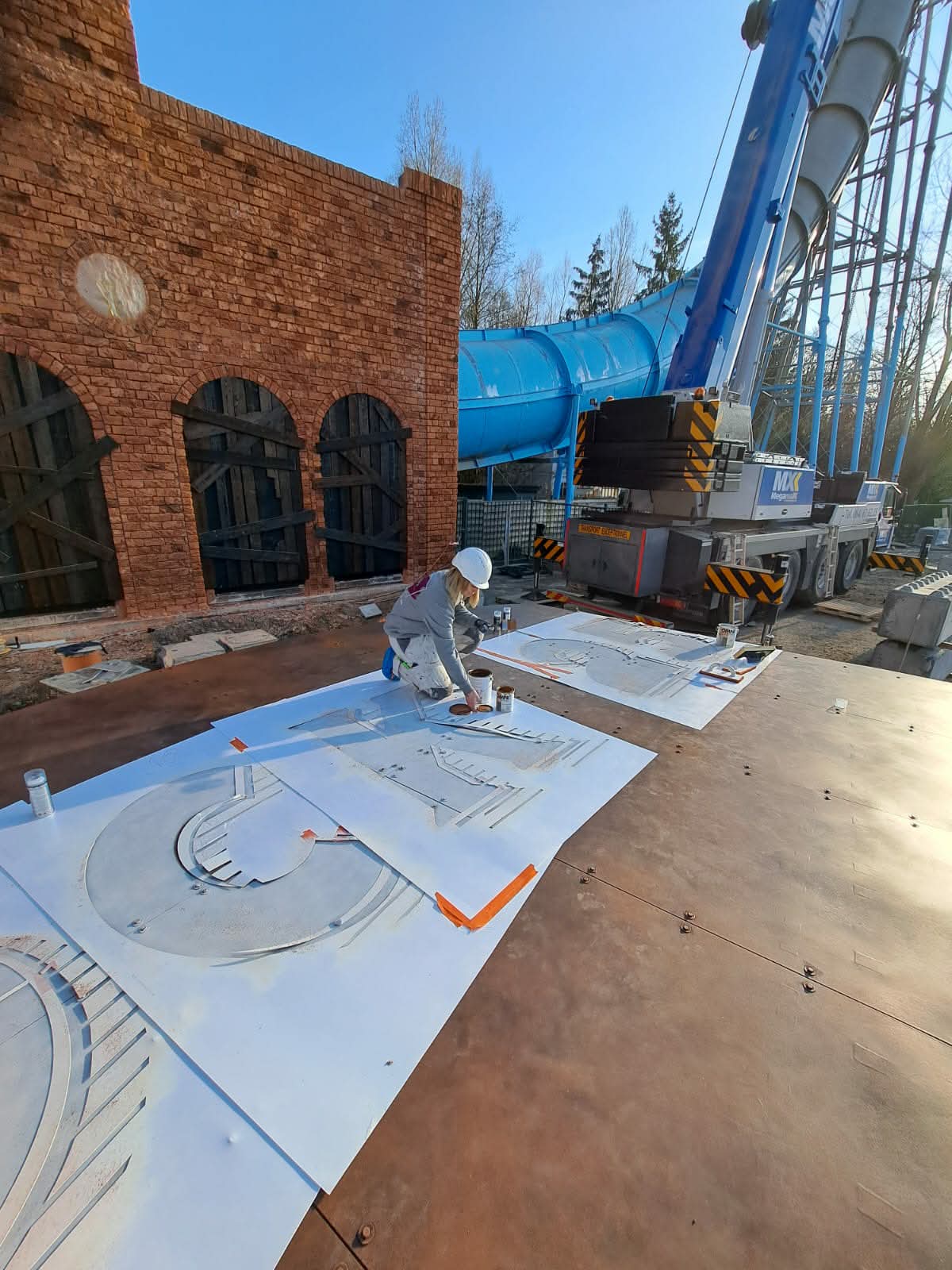 Ouvrière utilisant des pochoirs sur un chantier avec une grue et un toboggan bleu.